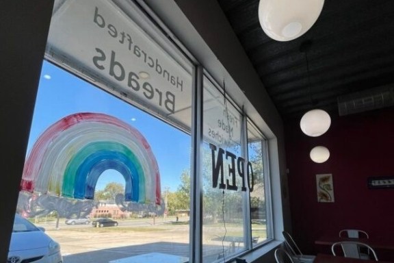 Cafe interior with large window, painted rainbow, 'OPEN' sign, and hanging lights.
