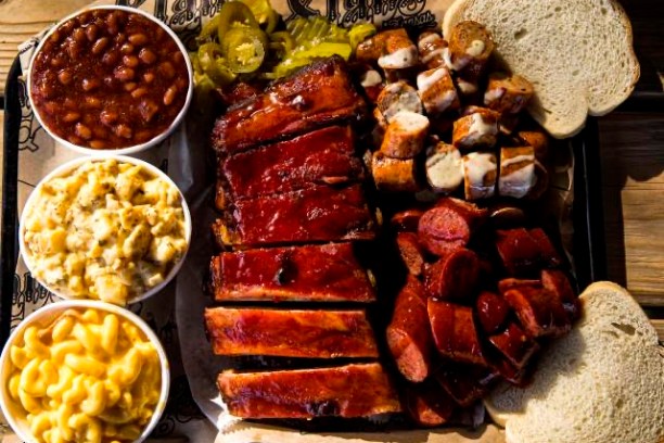 Barbecue platter with ribs, sausage, beans, mac and cheese, pickles, bread on wooden table.