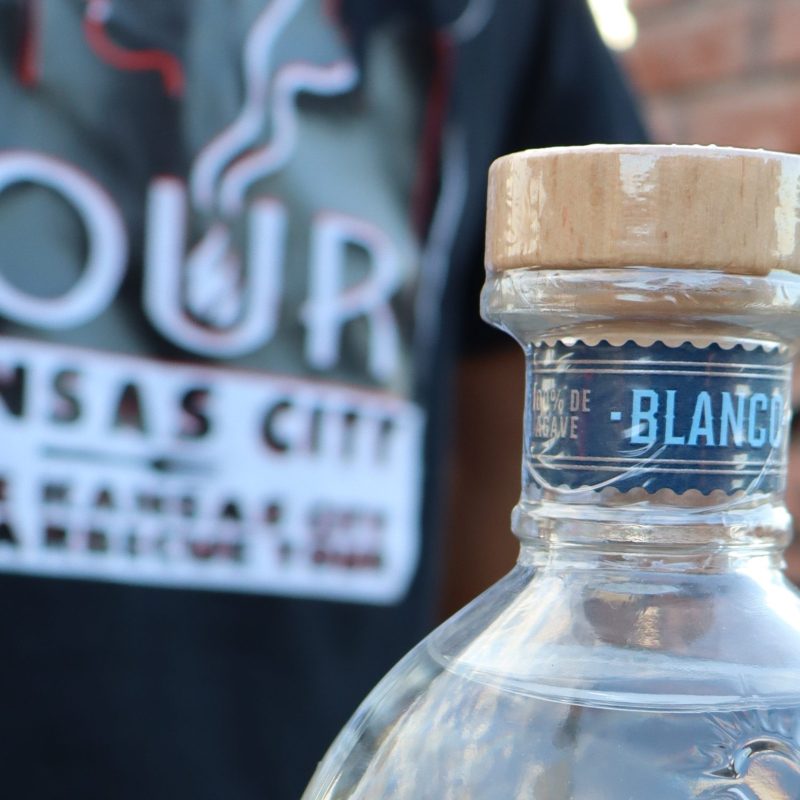 Astral Tequila Blanco bottle on a metal table, with a Kansas City tour T-shirt in the background.