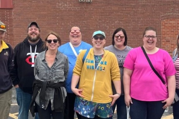 Group of seven adults smiling outdoors in casual clothing against a brick wall.
