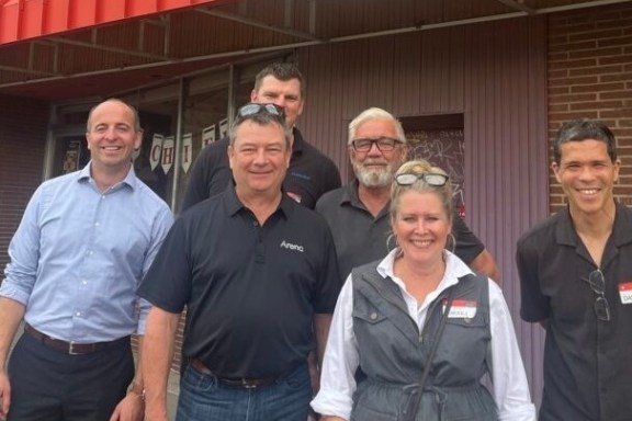 Six people smiling and standing outside a building with a red awning.
