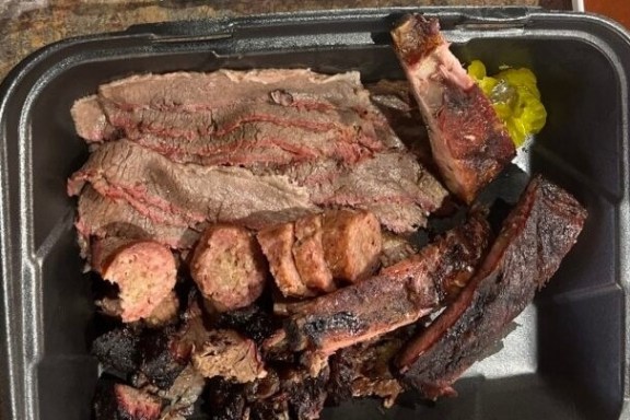 Assorted smoked meats including brisket, sausage, and ribs in a black takeout container.