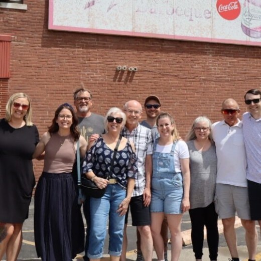 Group of 12 people posing in front of a brick wall with a faded Coca-Cola sign.