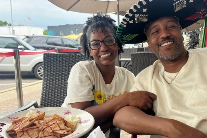 Smiling couple at outdoor cafe; man wears sombrero, plate of food and drink on table.