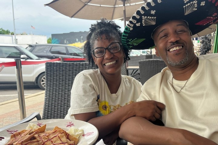 Smiling couple seated outdoors at a table with dessert and drink, one wearing a sombrero.