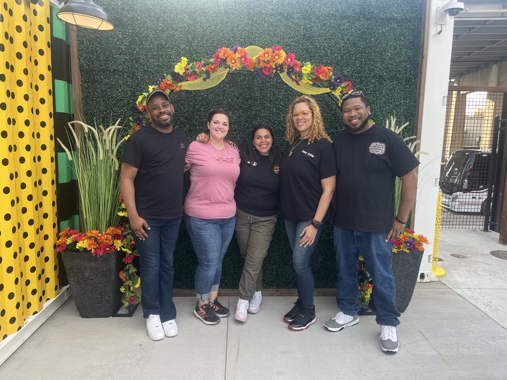 Five people smiling in front of a floral arch and green backdrop.