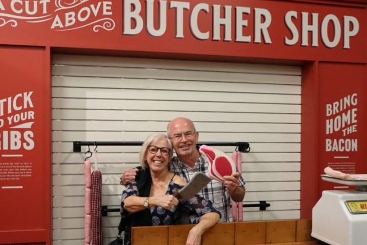 Two people smiling in a fake butcher shop holding a cleaver and meat props.