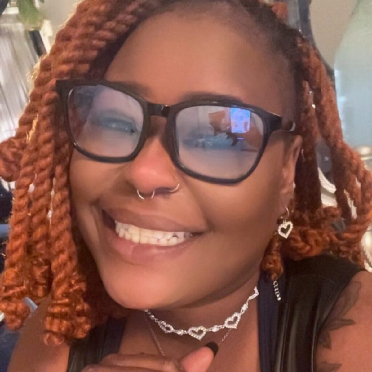 Smiling person with glasses and red braids, wearing heart jewelry and black nail polish.
