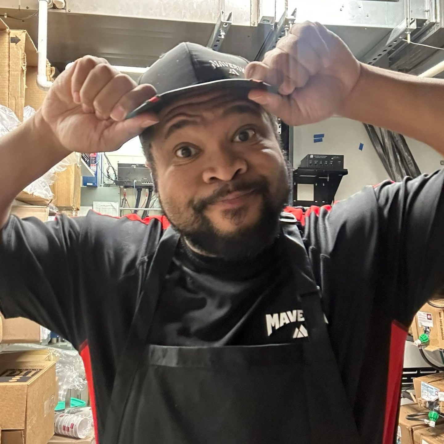 Man in a black apron and cap smiling, standing in an industrial storage area.