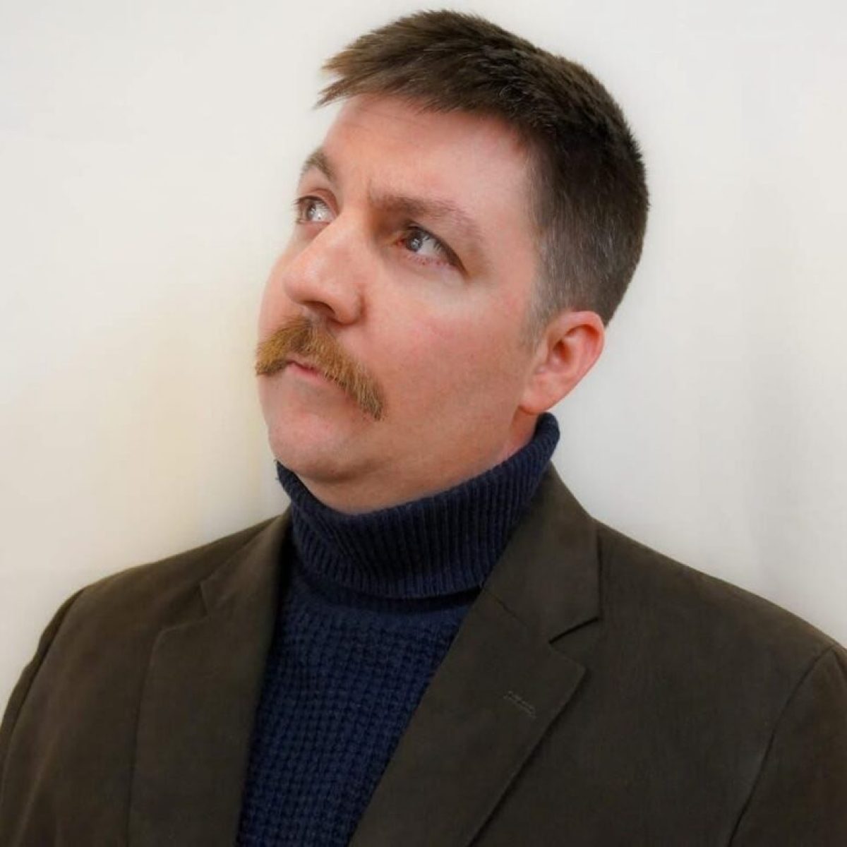 Person with short hair and mustache, wearing a turtleneck and blazer, looking upwards.