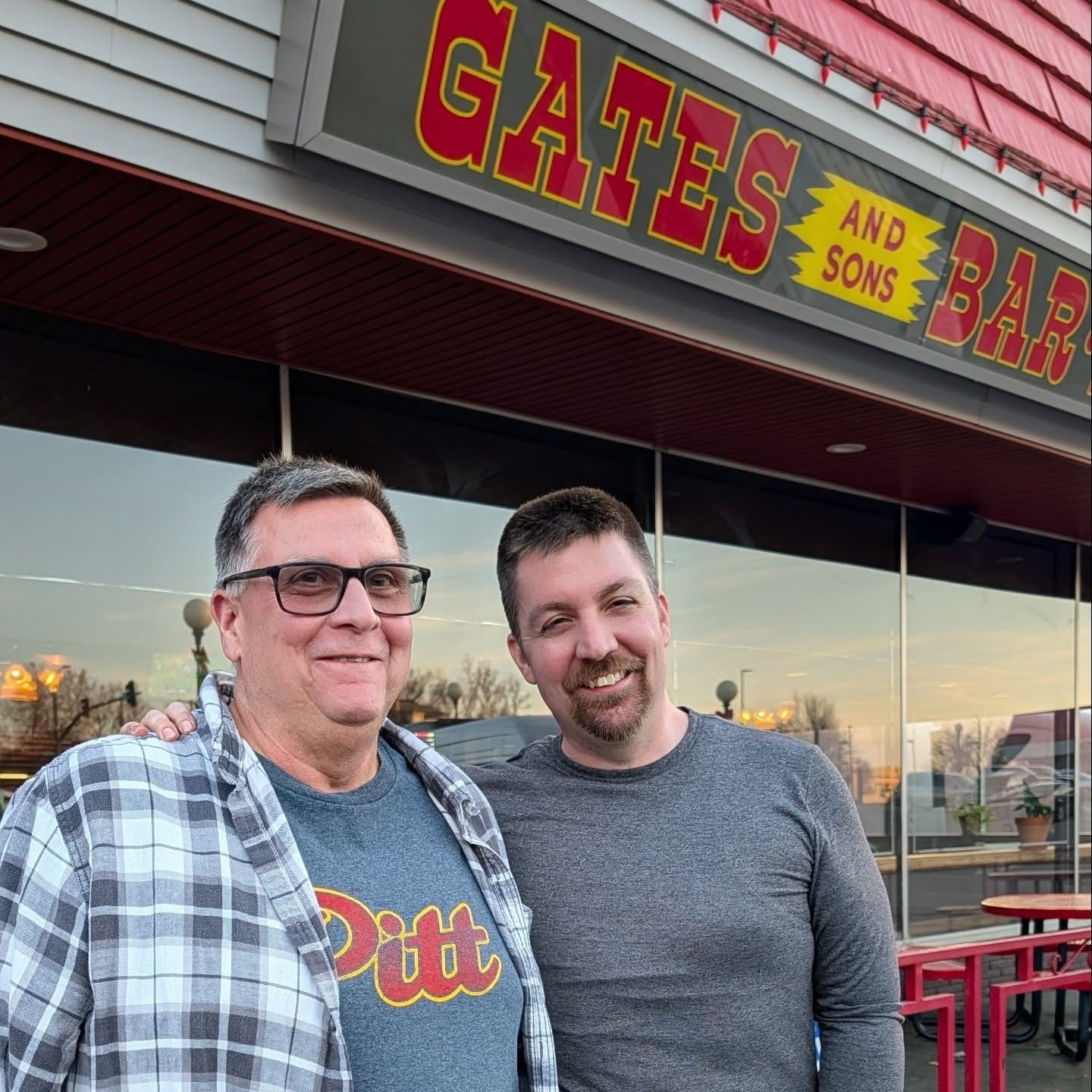 Two smiling men in front of Gates and Sons BBQ restaurant.