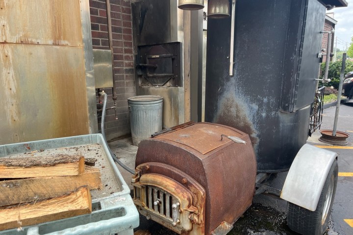 Outdoor smoker with rust, wood pile nearby, and metal trash can against brick wall.