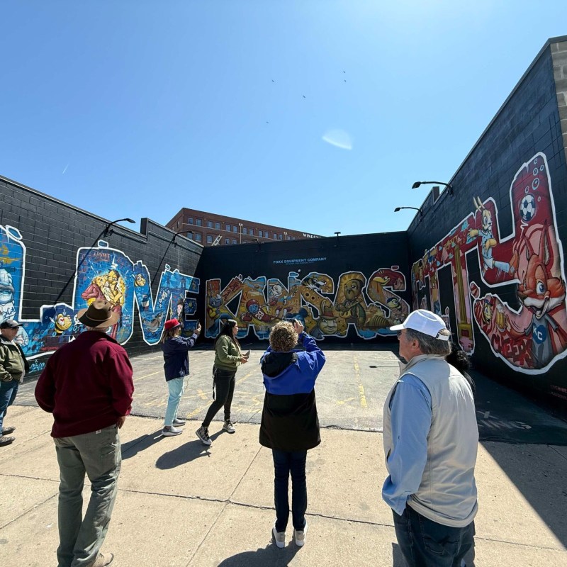 People observe a colorful mural saying 'Love Kansas City' on a sunny day.