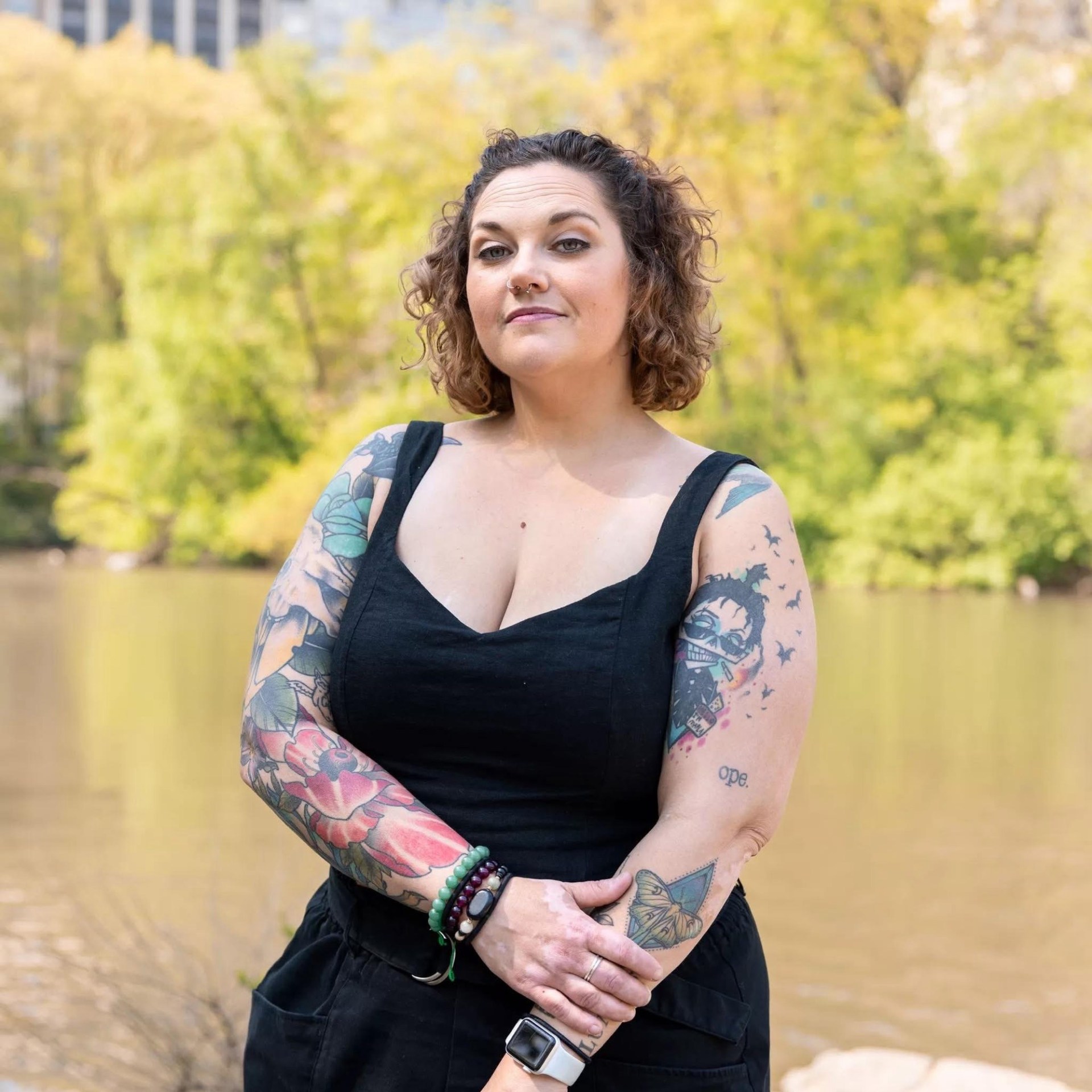 Person with tattoos, wearing black outfit, stands near water and trees.